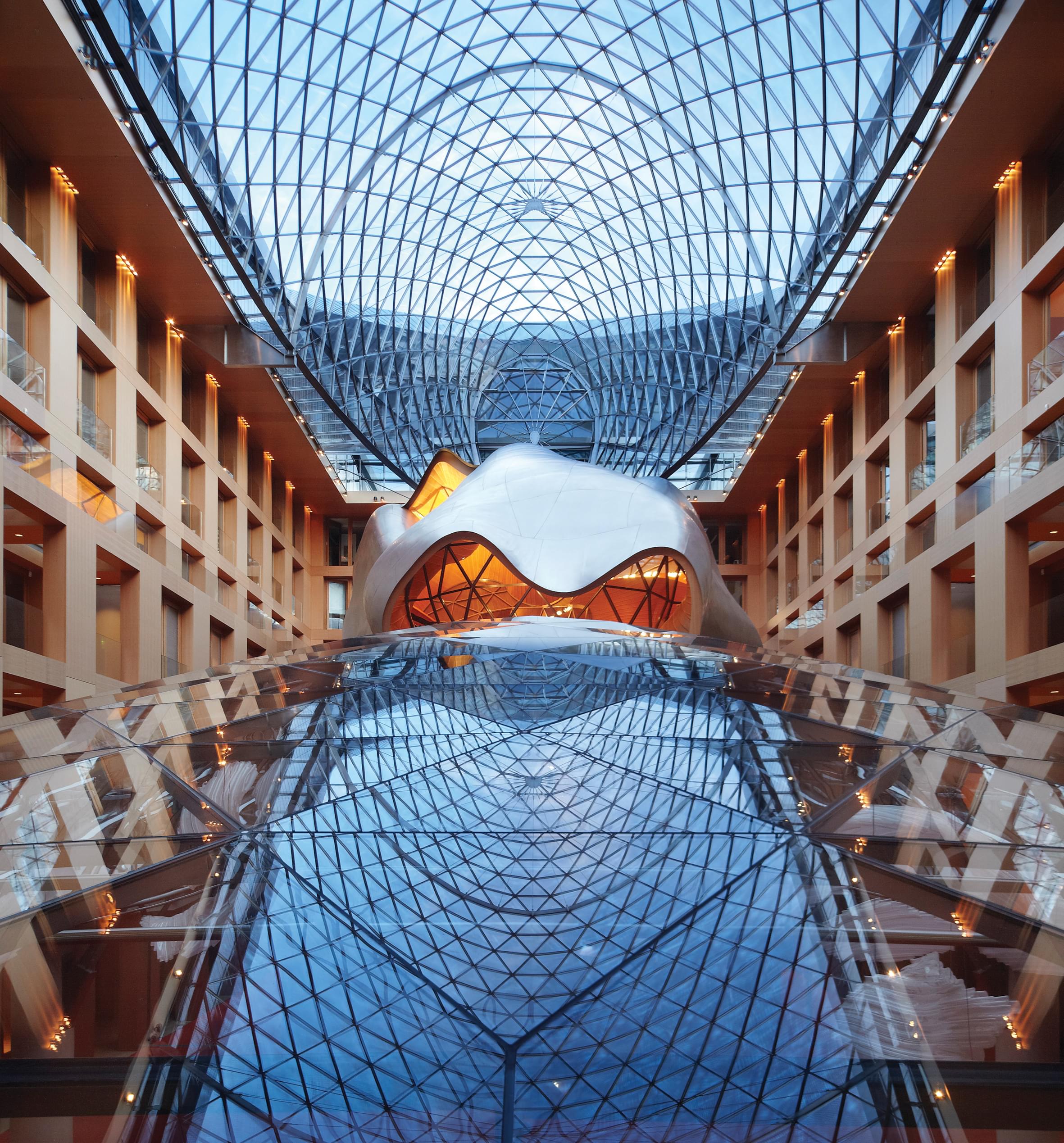 View into the atrium of the DZ BANK building: An inner courtyard emerges, surrounded by offices with pine wood façade and windows, above a vaulted glass roof and a steel sculpture in the centre of the courtyard. The vaulted glass roof of the basement rises in front of the sculpture.