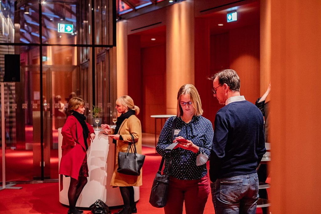 Guests at bar tables at a reception in the AXICA event location. The foyer with red carpet, surrounded by wooden pine panels, can be seen.